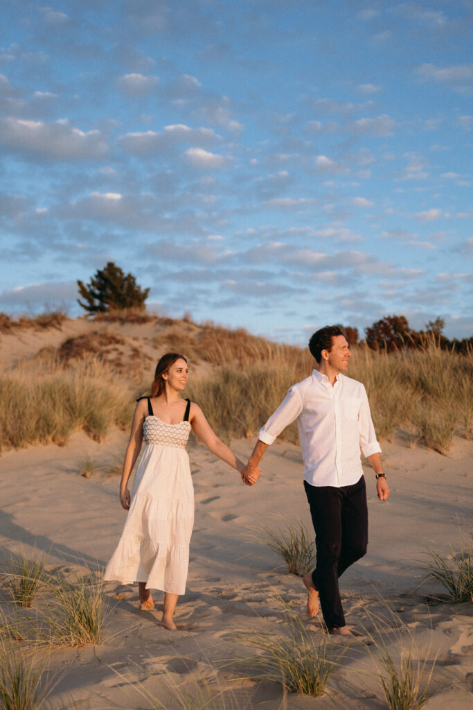 A couple walks hand in hand on a sandy beach at sunset, both barefoot. The woman wears a white dress, and the man is in a white shirt and dark pants. They are surrounded by sand dunes and tufts of grass under a partly cloudy sky.