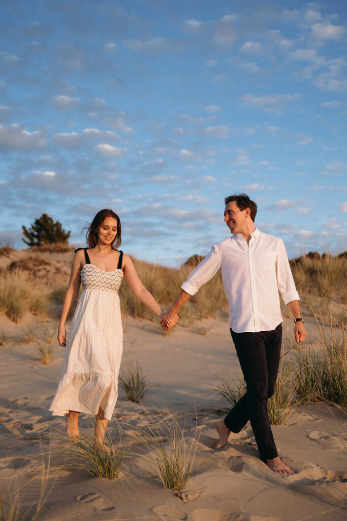 A couple walks hand in hand on a sandy beach at sunset. The woman is wearing a white dress and the man is in a white shirt and dark pants. They both appear happy, with grassy dunes and a cloudy sky in the background.