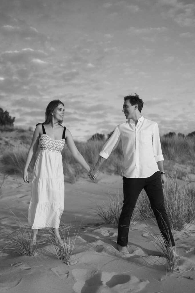 A black and white photo of a couple walking hand in hand on a sandy beach. The woman is wearing a long white dress and the man is in a white shirt and dark pants. They are smiling at each other under a partly cloudy sky.