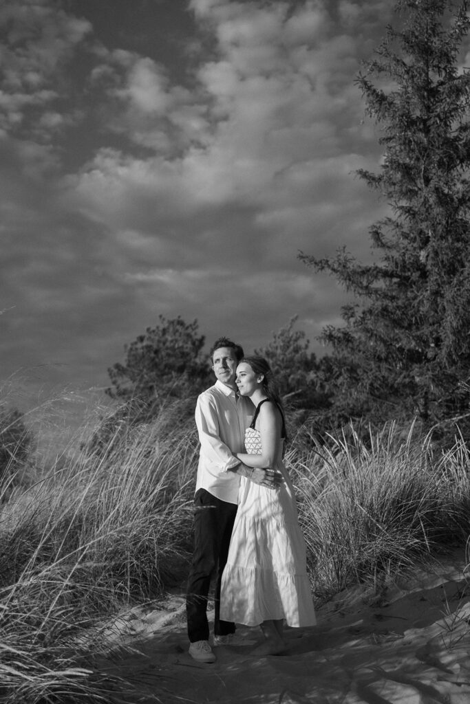 A couple stands in a grassy dune area, embracing. The woman wears a long dress, and the man wears a shirt and pants. Trees are in the background under a cloudy sky. The image is in black and white.