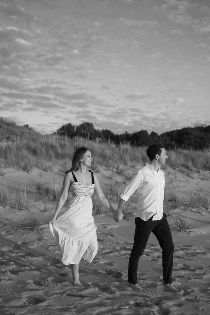 A couple holding hands walks barefoot on a sandy beach. The woman wears a long white dress, and the man wears a white shirt and dark pants. Grass-covered dunes and a partly cloudy sky are in the background. The image is in black and white.