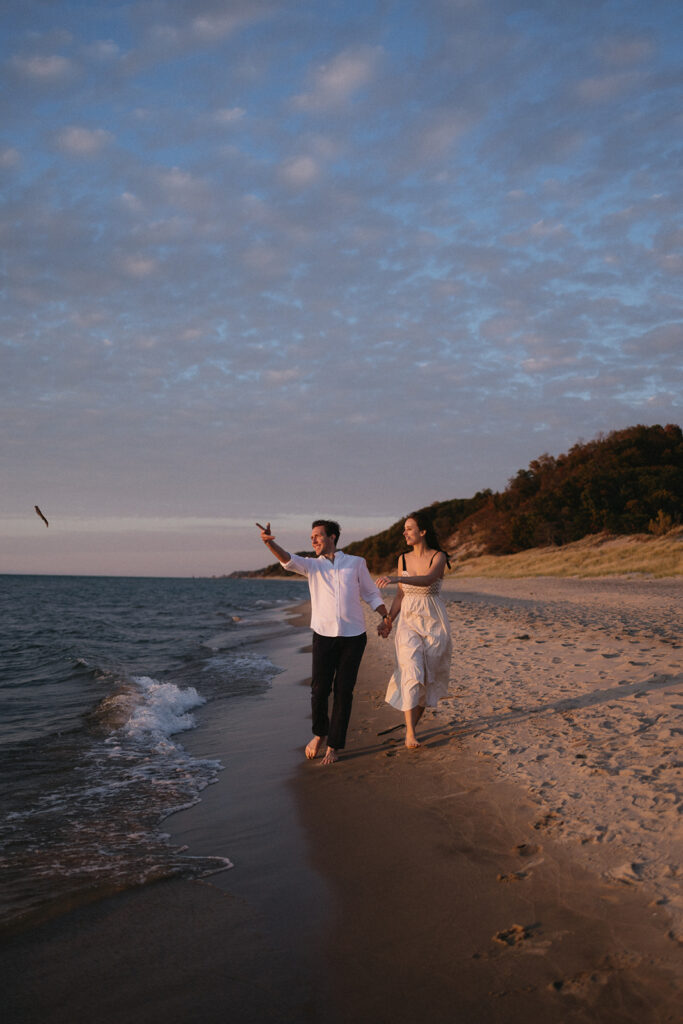 A couple walks barefoot along a sandy beach at sunset. The man in a white shirt throws a stick, while the woman in a white dress smiles. The ocean waves gently lap at the shore, and a hillside is visible in the background.