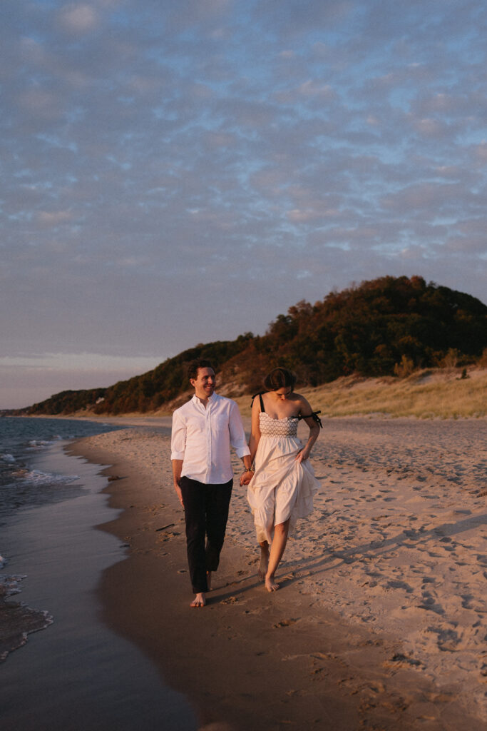 A couple holds hands while walking on a sandy beach at sunset. The man wears a white shirt and dark pants, and the woman wears a light dress. The sky is partly cloudy, and there are green hills in the background.