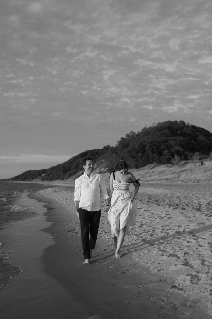 A couple walks barefoot along a sandy beach, holding hands. The man wears a light shirt and pants, while the woman wears a flowing dress. The sun is low, casting soft light, and a hill with trees is visible in the background. The sky has scattered clouds.