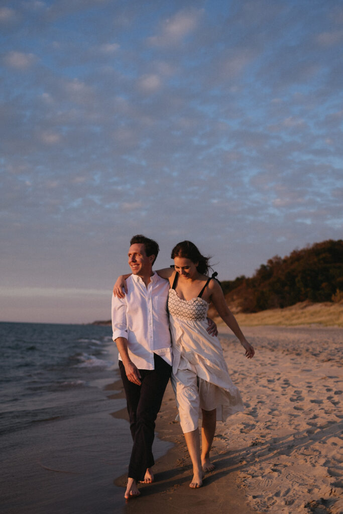 A couple walks barefoot along a sandy beach at sunset, with the ocean on their left. They are smiling, with the mans arm around the womans shoulders. The sky is partly cloudy, and the shoreline is lined with trees.