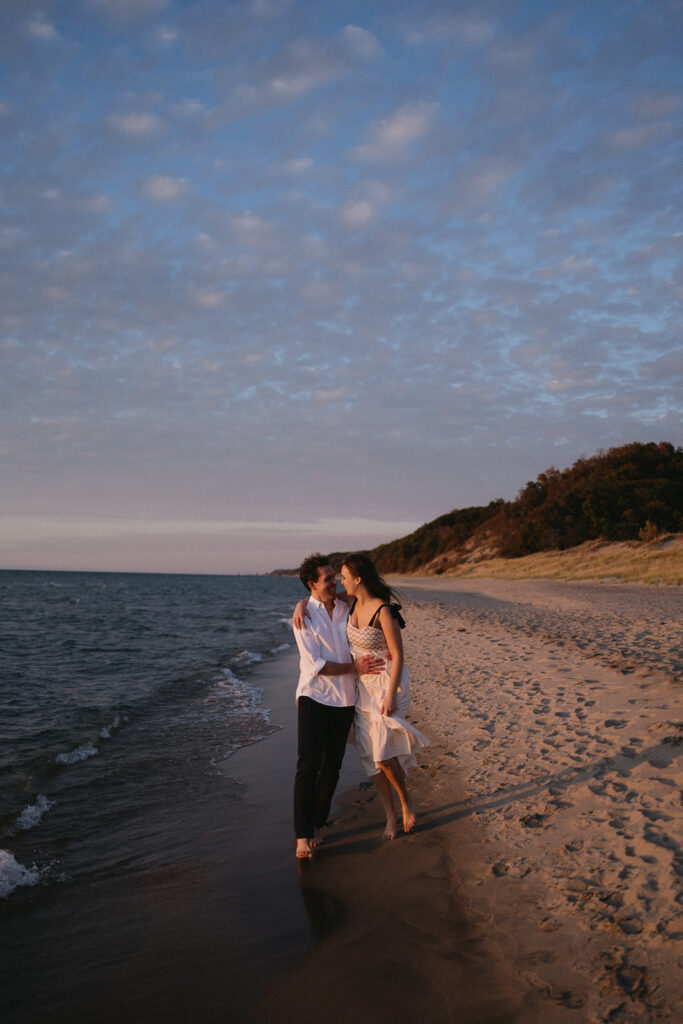 A couple walks barefoot along a sandy beach at sunset, with gentle waves lapping at their feet. They are smiling at each other, enjoying the calm ambiance. The sky is partly cloudy, and there are hills in the background.