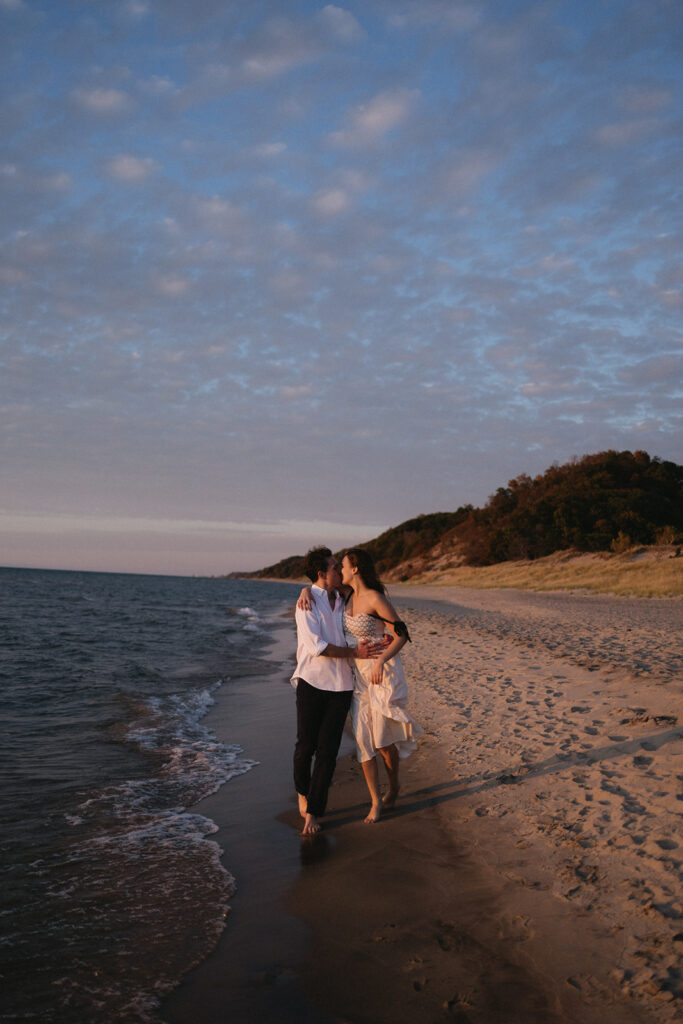 A couple walks hand-in-hand along a sandy beach at sunset. The man wears a white shirt and dark pants, and the woman wears a flowing dress. Waves gently lap at the shore, and the sky is dotted with soft clouds with hints of pink and orange.