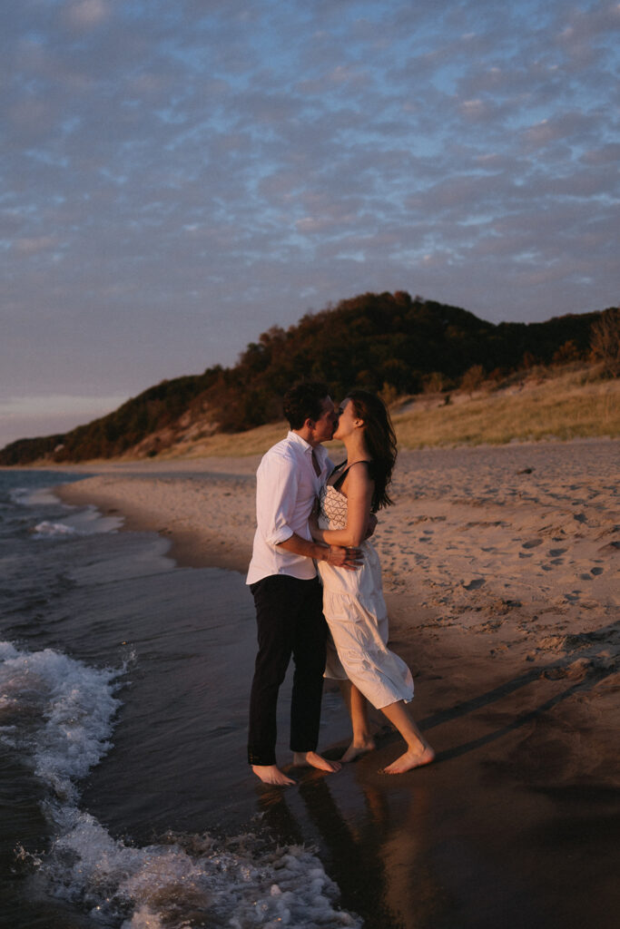 A couple stands on a sandy beach at sunset, embracing and smiling. The waves gently lap at their feet. The sky is dotted with clouds, and a hillside covered in greenery is visible in the background.