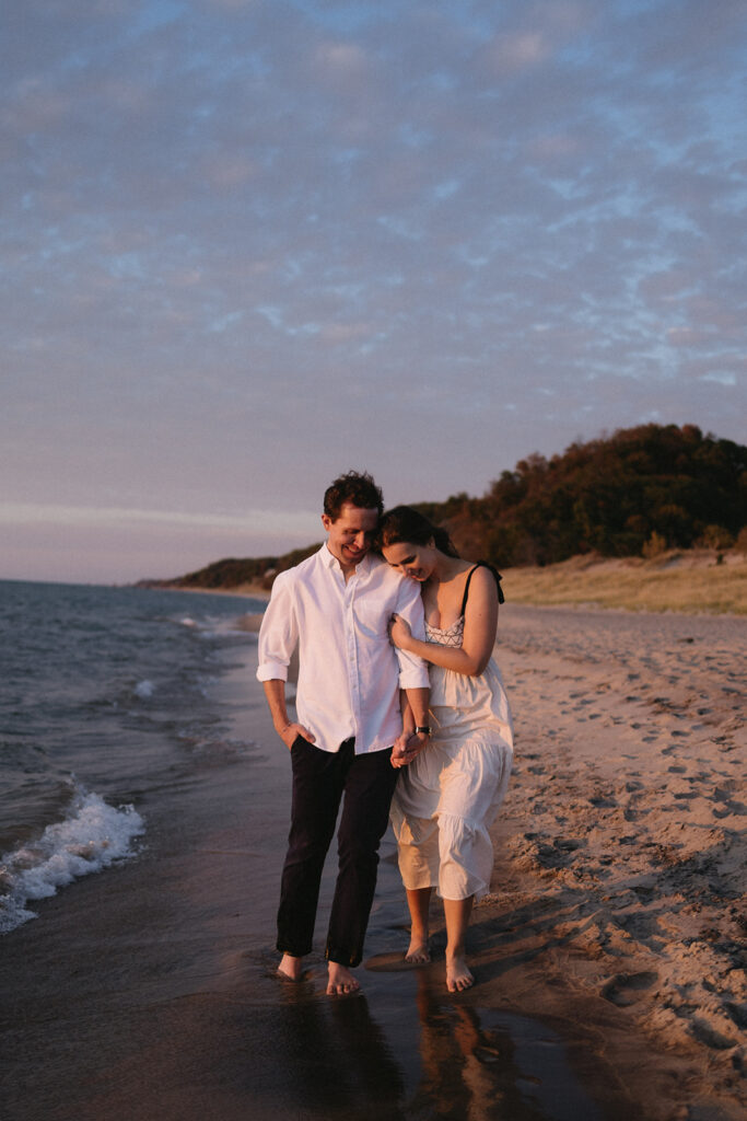 A couple walks barefoot along a sandy beach at sunset, holding hands. The man wears a white shirt and dark pants, while the woman wears a light dress. The sky is partly cloudy, and the ocean is gently lapping at the shore.