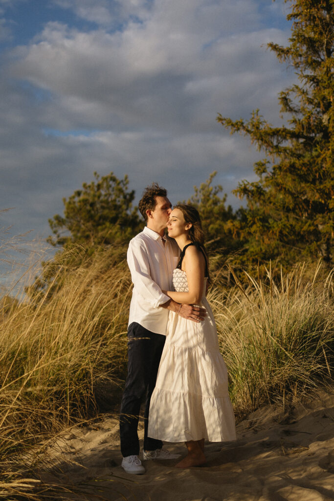 A couple stands on a sandy path surrounded by tall grass. The man, wearing a white shirt and dark pants, embraces the woman, who is wearing a white dress. They are under a cloudy blue sky with trees in the background.