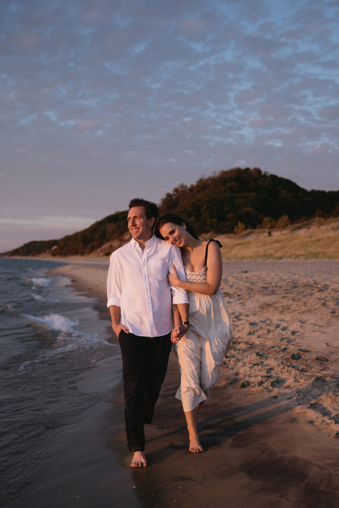 A couple walks barefoot along a sandy beach during sunset. The man wears a white shirt and dark pants, while the woman in a white dress leans on him. They are smiling, with a grassy hill in the background and the ocean to their side.