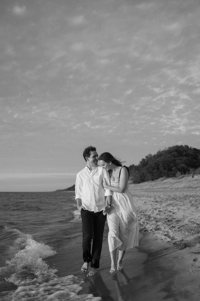 A couple walks barefoot along a sandy beach, holding hands and smiling. The woman wears a light dress and the man is in a shirt and pants. Gentle waves lap at their feet under a sky filled with scattered clouds. The scene is in black and white.