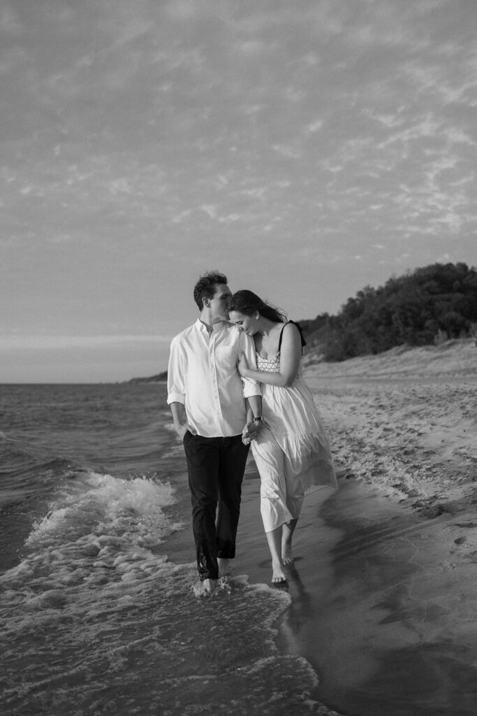 A black and white photo of a couple walking barefoot on a beach. The man is wearing a white shirt and dark pants, and the woman is in a light dress. They are holding hands, with gentle waves lapping at their feet. Trees line the shore in the background.