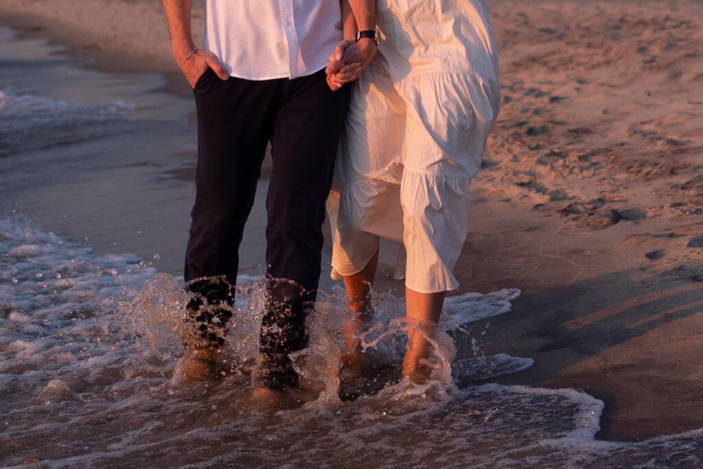 A couple walking hand in hand along a sandy beach at sunset. Water splashes around their feet; the man is in dark pants and a white shirt, and the woman is in a white dress.