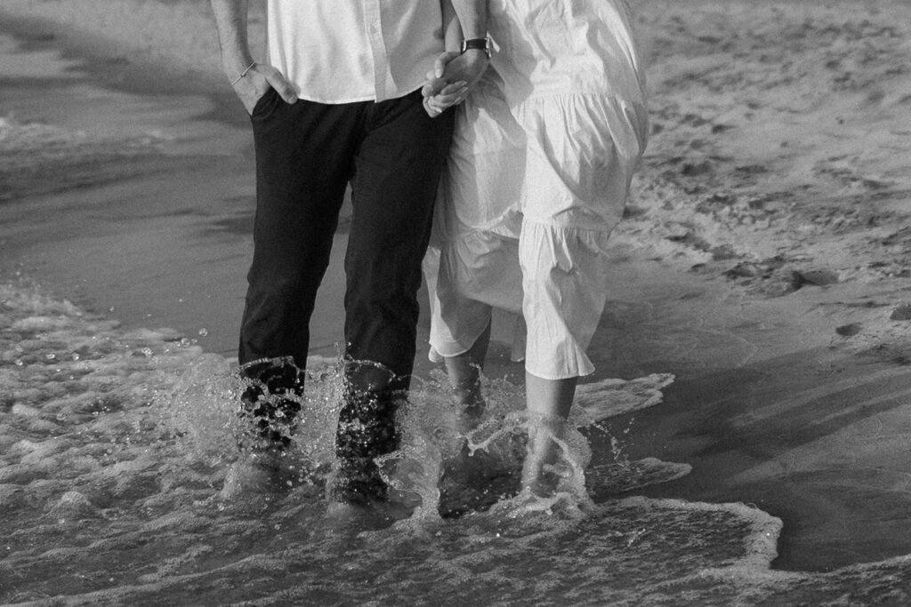 A couple walks along a beach with water splashing around their legs. They hold hands, and the person on the left wears dark pants while the person on the right wears a white dress. The scene is in black and white.