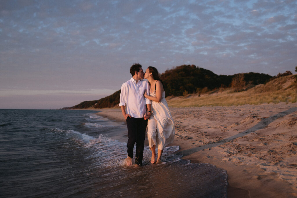 A couple walks hand in hand along a beach at sunset. The woman in a white dress and the man in a white shirt are barefoot, enjoying the waves. The sky is partly cloudy with hues of pink and orange, and there are dunes and greenery in the background.