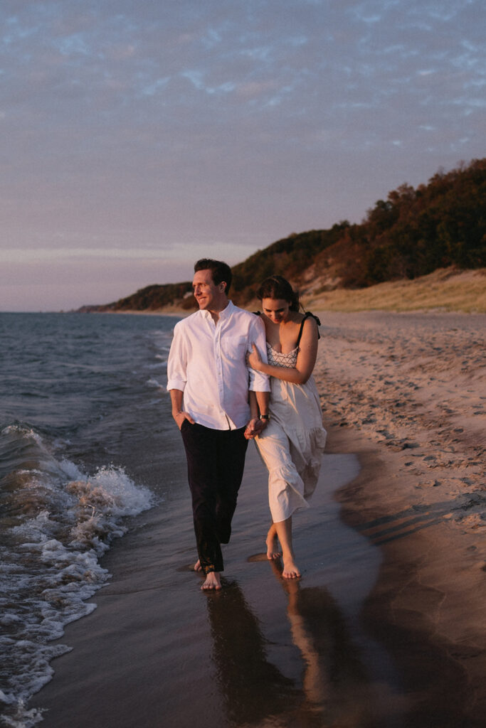 A couple walks barefoot along a sandy beach at sunset. The man wears a white shirt and dark pants, while the woman wears a white dress, holding his arm. The sky is painted with soft purple and orange hues, and gentle waves touch the shoreline.