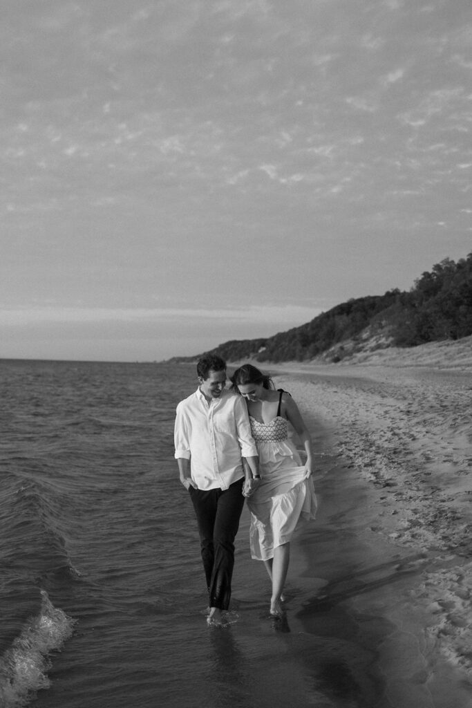 A black and white image of a couple walking hand in hand along the shoreline. They are both smiling, with the woman in a light dress and the man in a white shirt and dark pants. The beach is mostly empty, with gentle waves and distant trees visible.