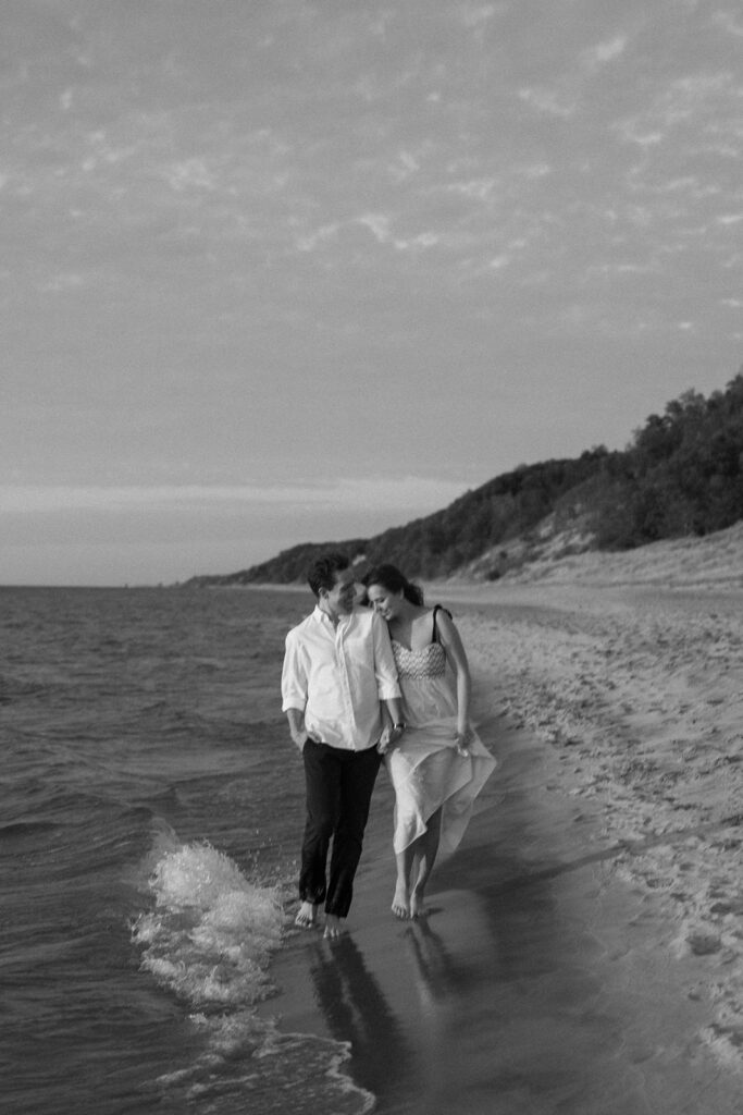 A black and white photo of a couple walking barefoot along a beach shoreline, holding hands. The man wears a white shirt and dark pants; the woman wears a light dress. Waves gently lap at their feet, and a wooded hill is visible in the background.