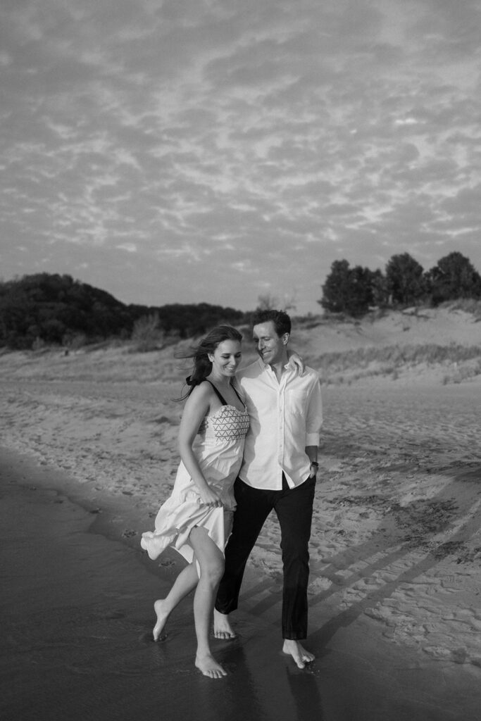 A couple walks barefoot along a sandy beach, with gentle waves lapping at their feet. The woman wears a white dress, and the man wears a white shirt and dark pants. The sky is cloudy, and dunes with greenery are in the background. The image is in black and white.