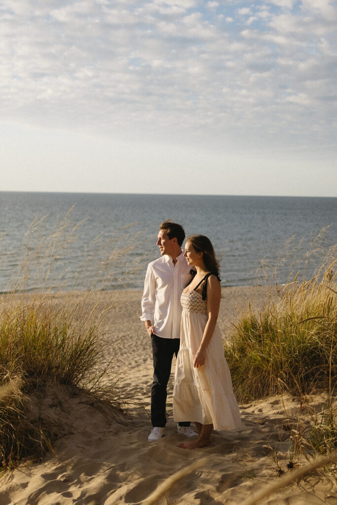 A couple stands together on a sandy beach, surrounded by dune grass. They are looking into the distance, enjoying a serene moment. The man wears a white shirt and dark pants, and the woman is in a light dress. The ocean and a partly cloudy sky are in the background.