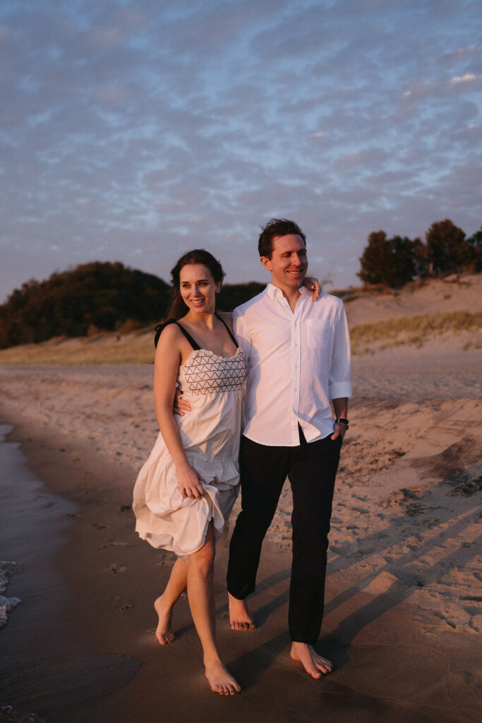 A couple walks along a beach at sunset. The woman wears a white dress, and the man wears a white shirt and dark pants. They appear happy and relaxed, with the ocean and a distant tree line in the background.