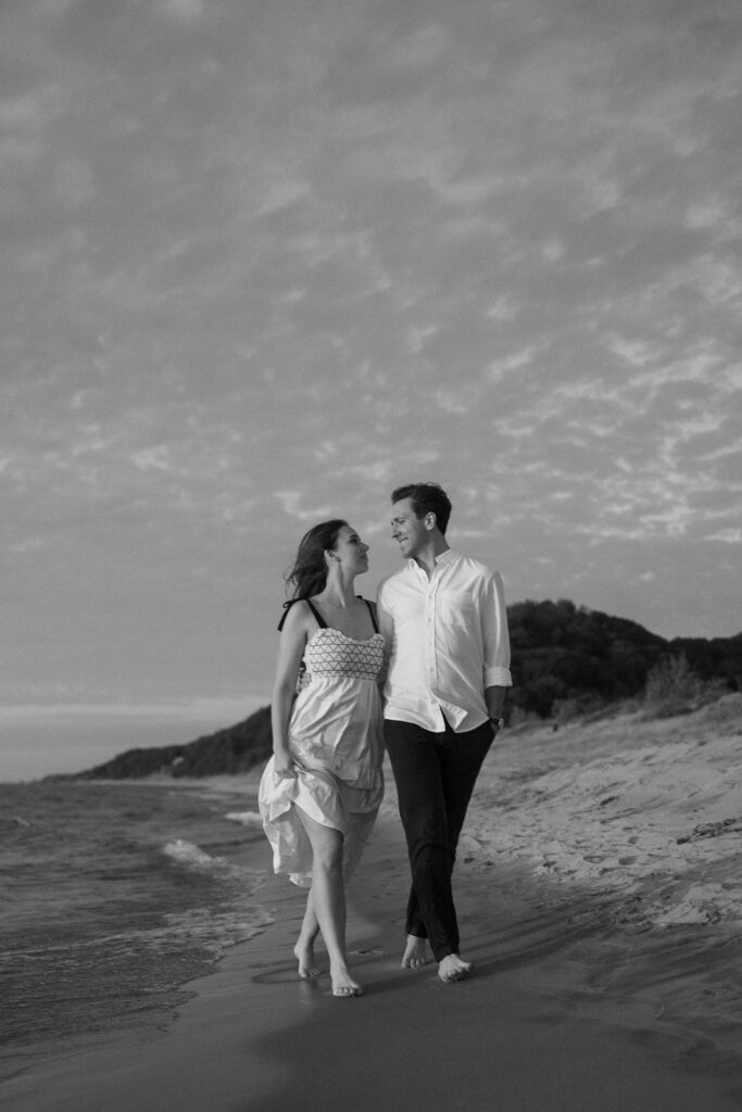 A couple walks barefoot along a sandy beach at sunset. The woman wears a light dress, and the man is in a white shirt and dark pants. They are smiling at each other, with waves gently lapping at the shoreline in the background.