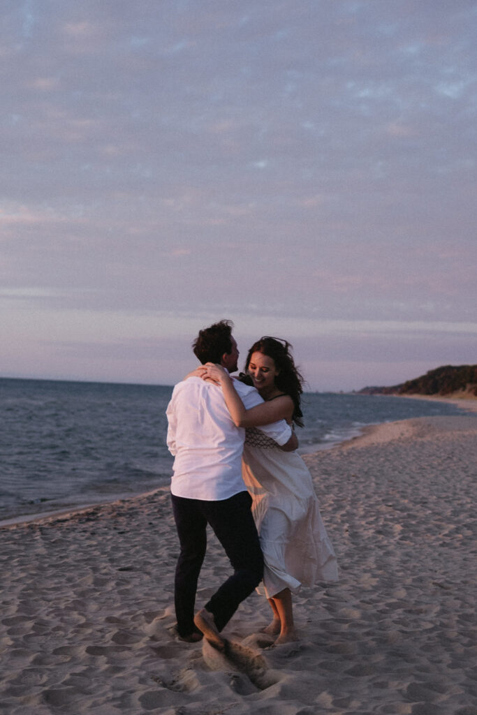 A couple dances barefoot on a sandy beach at sunset. The woman is wearing a flowing white dress, and the man is in a white shirt and dark pants. The sky is a mix of pink and purple hues, and the ocean is calm beside them.