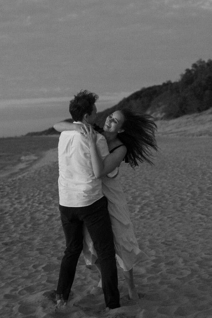 A black and white photo of a couple embracing and laughing on a beach. The woman has long hair and is wearing a dress, while the man is in a shirt and pants. The beach is expansive with a distant treeline under a cloudy sky.