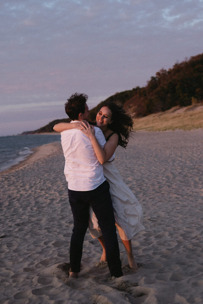 A couple dances joyfully on a sandy beach during sunset. The woman, wearing a white dress, smiles at the man in a white shirt and dark pants. The sky is tinged with pink and purple hues, and a tree-lined hill is visible in the background.