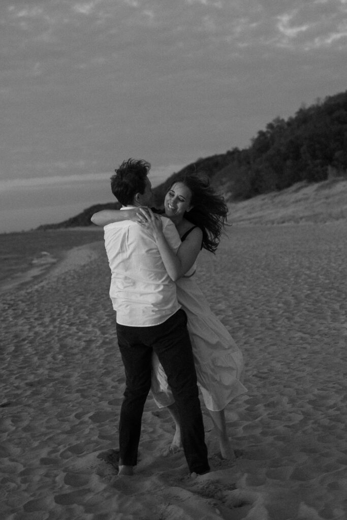 A black-and-white photo of a couple laughing and embracing on a sandy beach. The woman is wearing a light dress, and the man is in a white shirt and dark pants. The shoreline and a hillside with trees are in the background. The scene is joyful and serene.