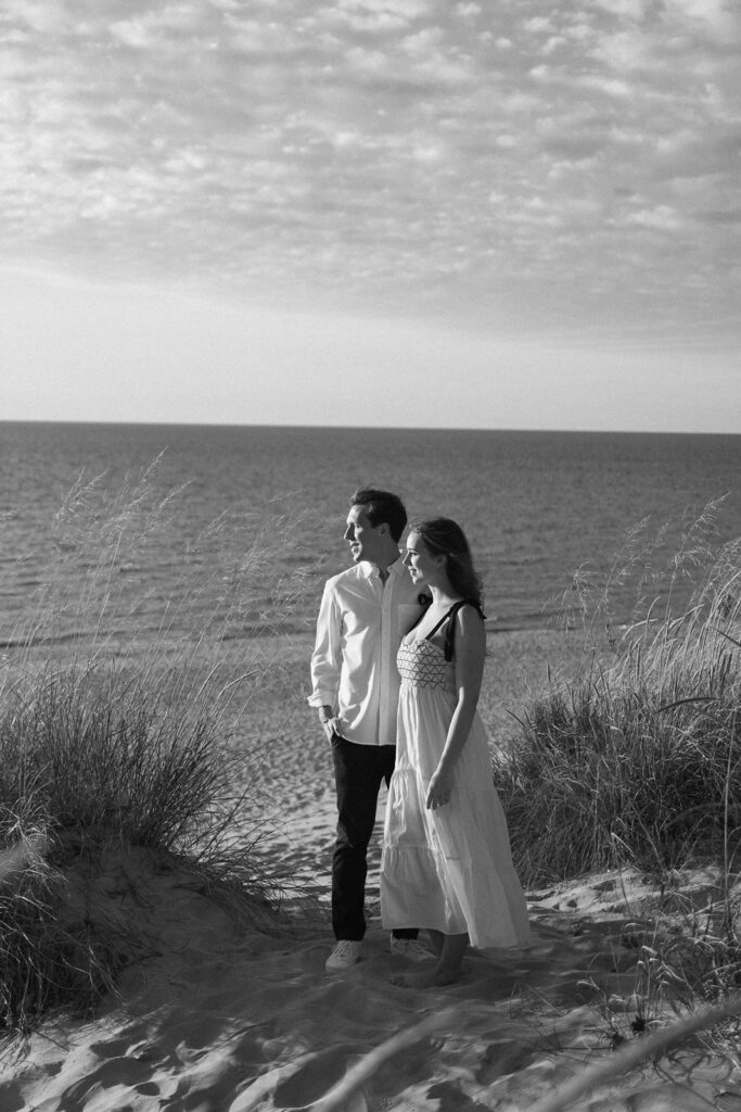 A black-and-white photo of a couple standing on a sandy beach path, gazing toward the ocean. The man wears a white shirt and dark pants, and the woman is in a long dress. Grasses frame the path under a partly cloudy sky.