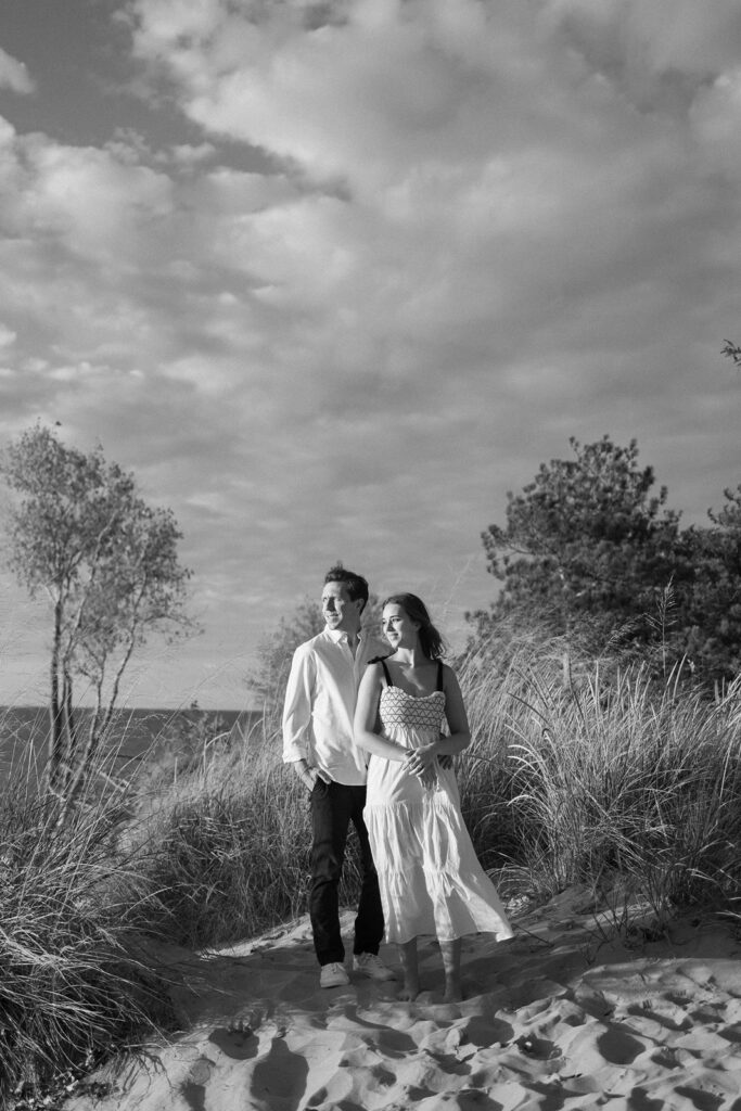 A couple walks hand in hand along a sandy path surrounded by tall grasses. The sky is partially cloudy, and the scene is in black and white, creating a serene and timeless atmosphere.