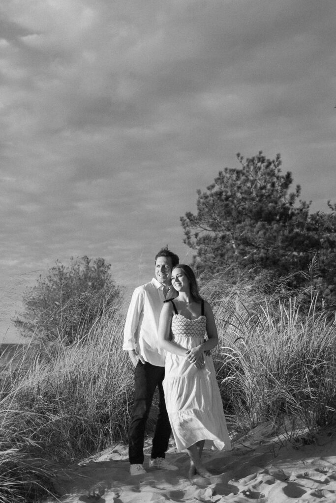 A black-and-white photo of a couple walking barefoot on a beach. The man is wearing a light shirt and dark pants, and the woman is in a light dress. They are smiling, surrounded by dune grasses and trees under a cloudy sky.