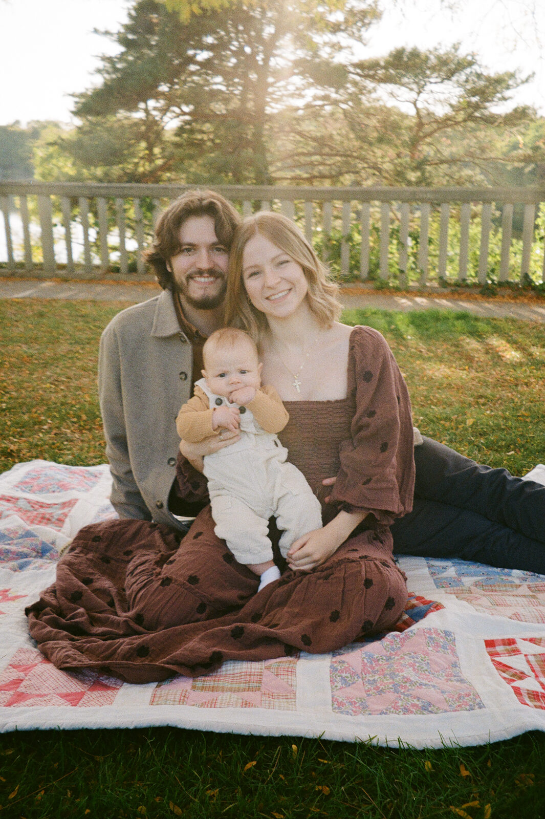 A family sits on a quilt in a grassy area. A man with long hair is wearing a gray coat. A woman with long blond hair is wearing a brown dress and holding a baby. The baby is dressed in a light outfit. Trees and a wooden fence are in the background.