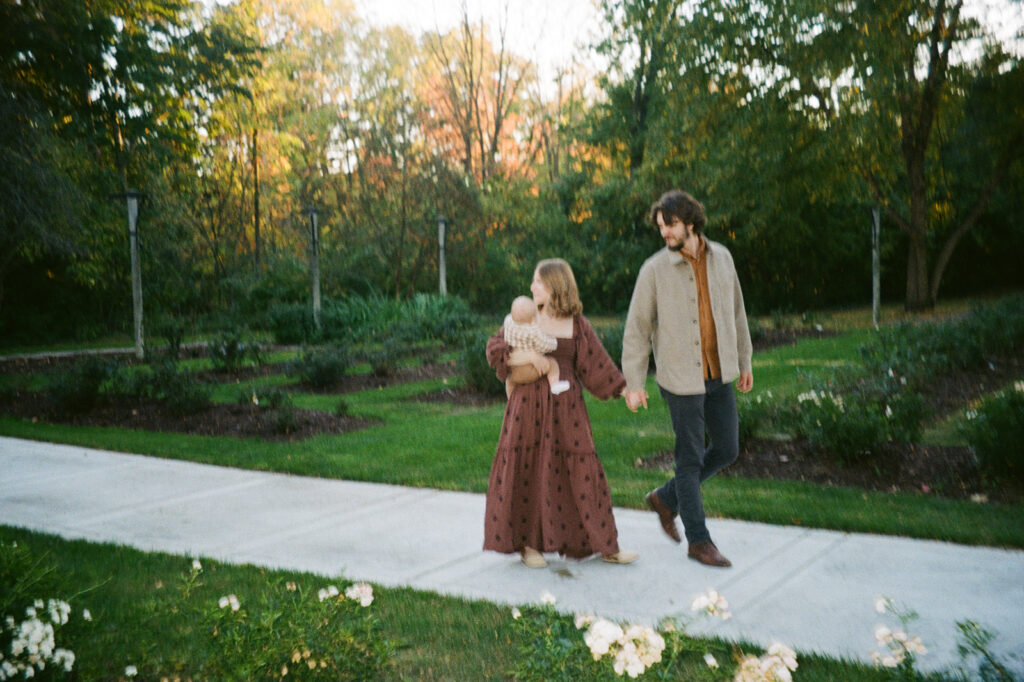 A couple walks hand in hand on a garden pathway. The woman, wearing a long patterned dress, holds a baby. The man wears a cardigan and trousers. The background features greenery and trees under a partly cloudy sky.