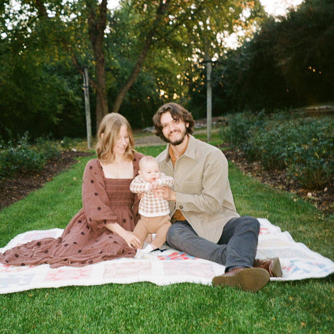 A couple sits on a picnic blanket in a garden, smiling at the camera. The woman holds a baby in her lap. They are surrounded by greenery and trees, with a path leading away behind them.