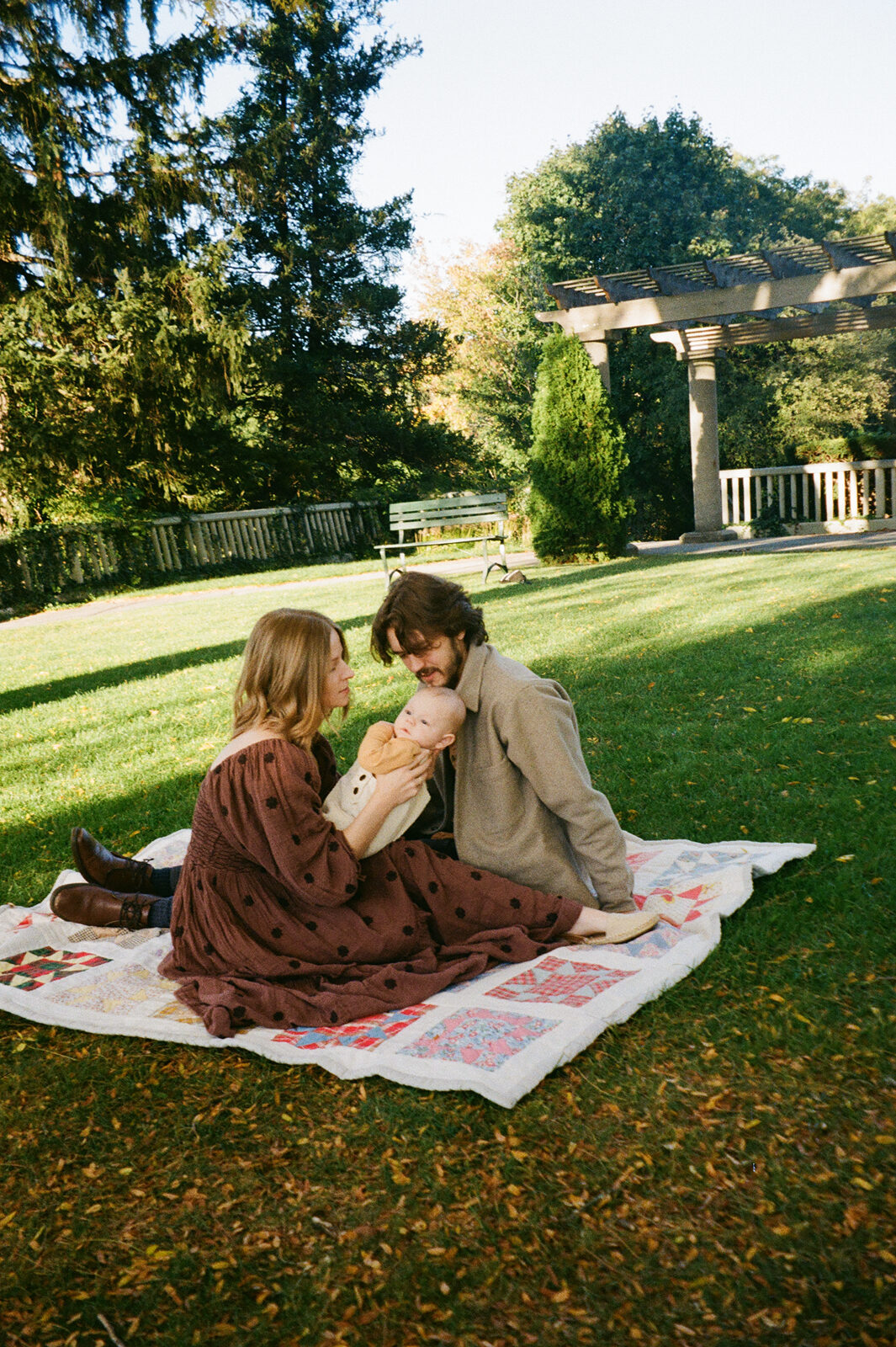 A couple sits on a patterned blanket on grass, facing each other. The woman holds a baby, and they share a tender moment. Trees, a wooden structure, and a sunny sky fill the background.