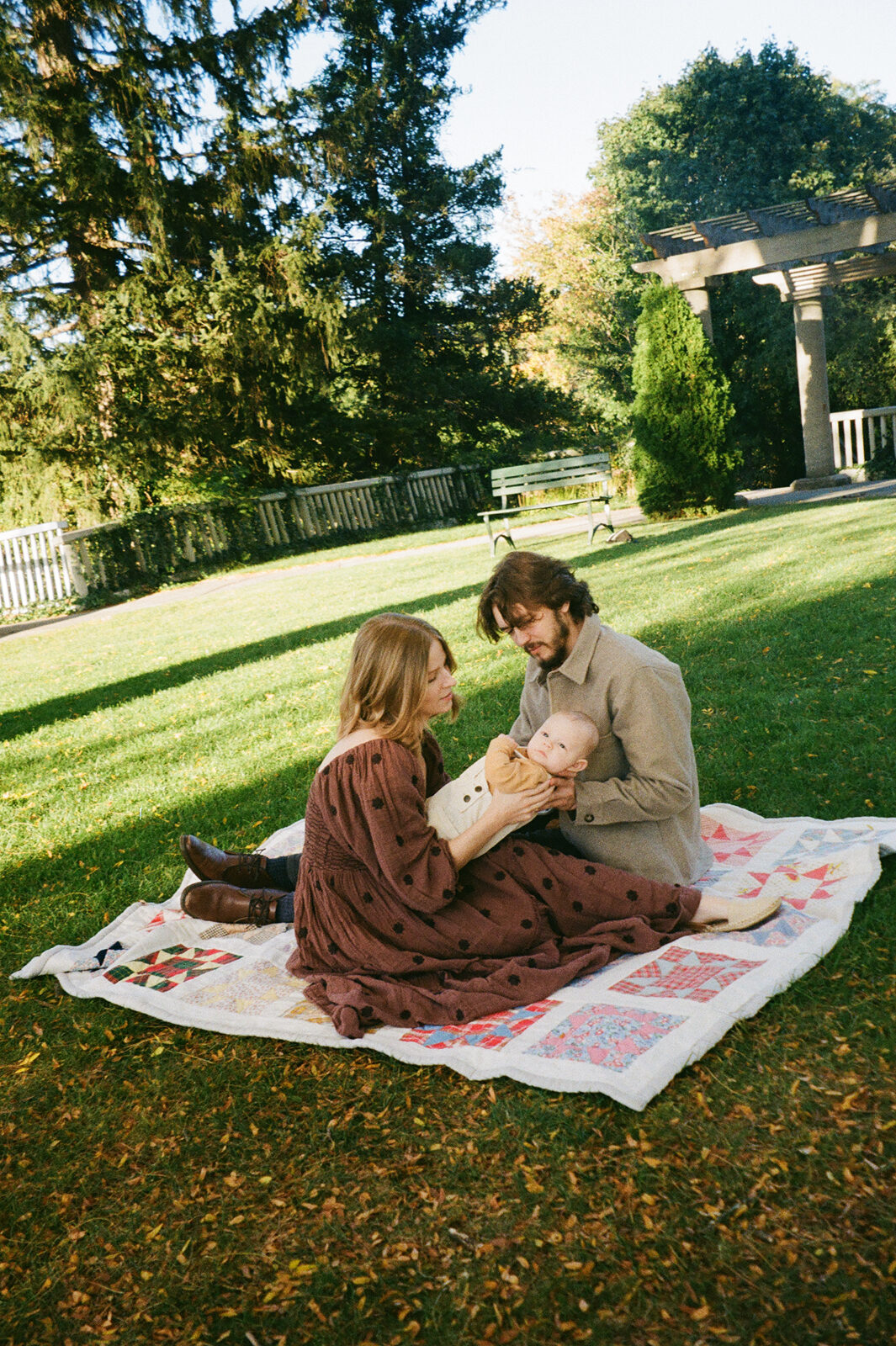 A couple sits on a quilt in a grassy park, playing with a baby. Theyre surrounded by trees and a pergola. The woman is wearing a brown coat, and the man is in a tan jacket. The sun casts shadows on the ground.