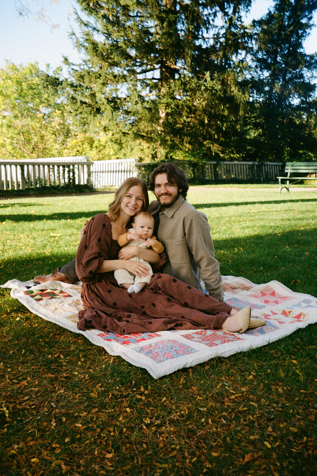 A couple sits on a patterned blanket in a grassy area, holding a baby. They are surrounded by trees and a wooden fence. The scene is sunny and serene, suggesting a peaceful outdoor family moment.