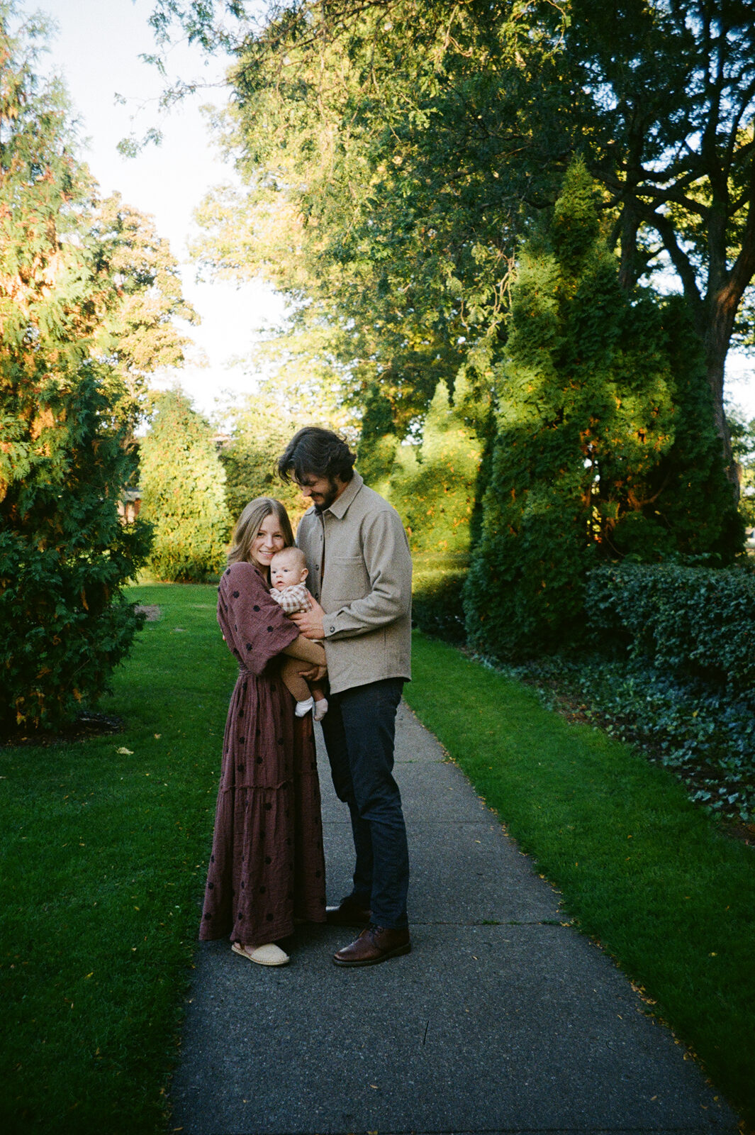 A couple embraces on a path in a garden. The woman wears a long brown dress, and the man is in a beige jacket and pants. The path is lined with grass and trees, bathed in sunlight.