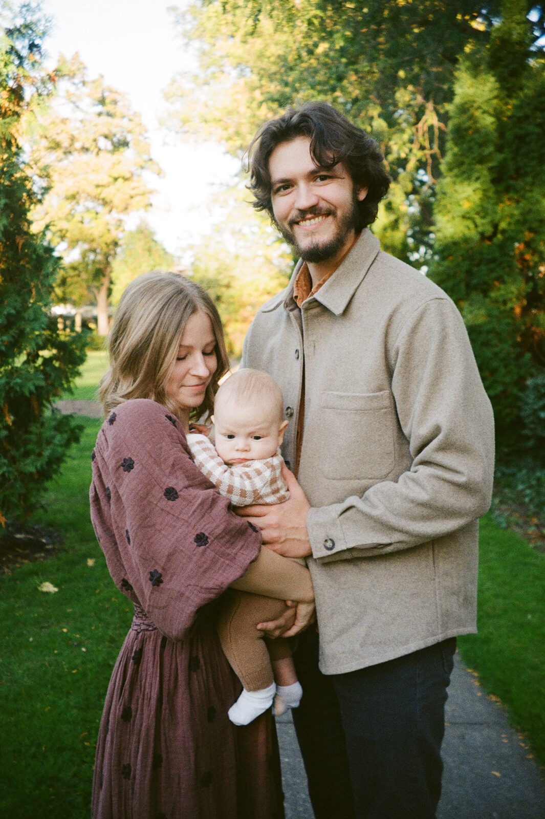 A couple stands outside on a sunny day, holding a baby. The woman, in a maroon dress, leans toward the baby, who wears a plaid shirt and brown pants. The man in a brown jacket smiles at the camera. Green trees and grass are in the background.