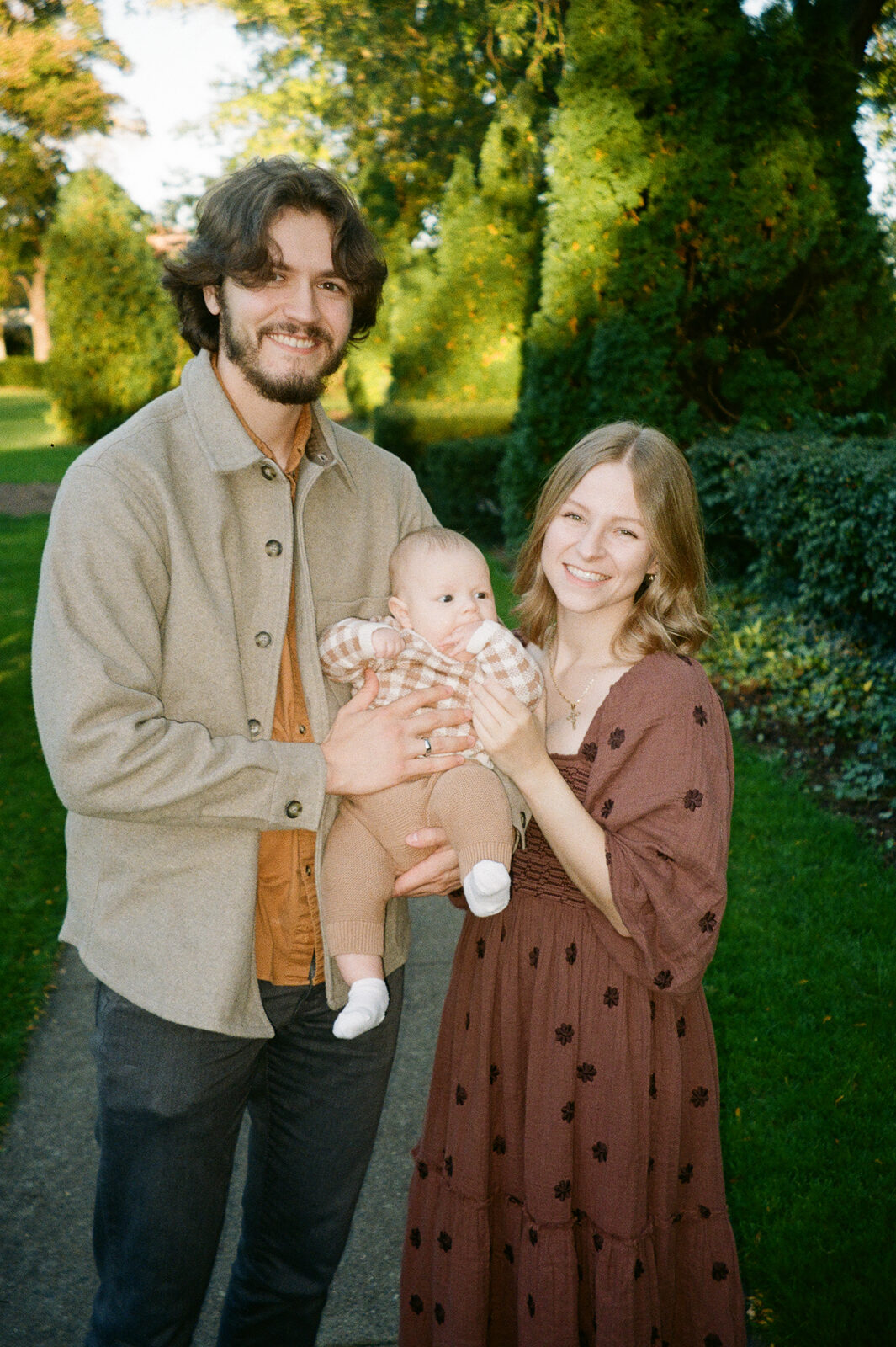 A couple is standing outdoors on a path surrounded by greenery, smiling at the camera. They are holding a baby dressed in a plaid outfit. The woman is wearing a brown dress, and the man is in a beige jacket with an orange shirt.