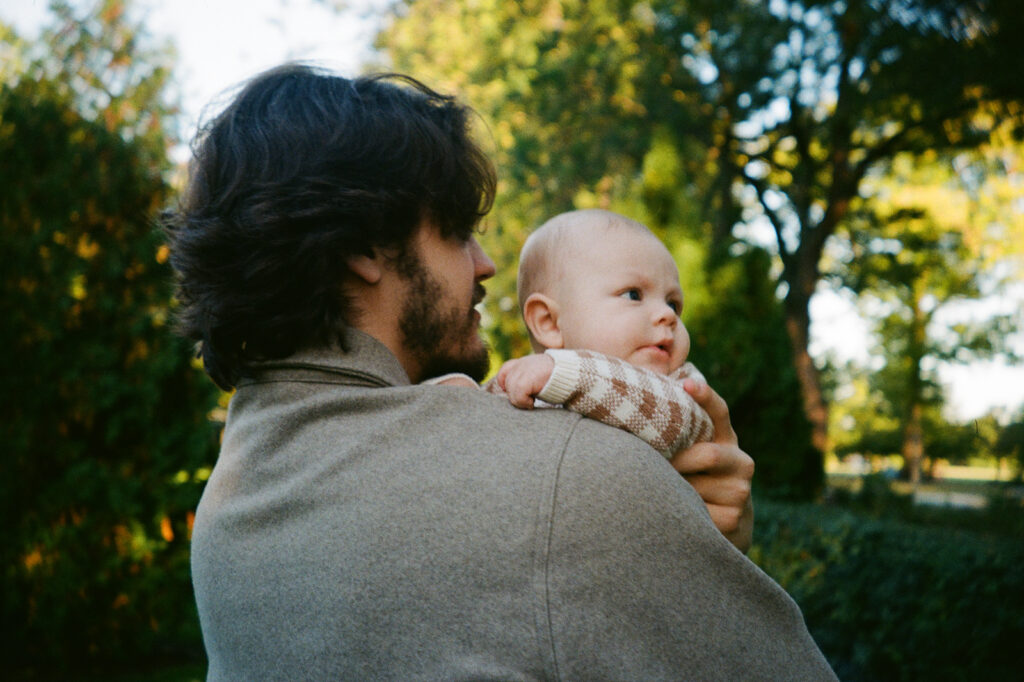 A man with dark hair holds a baby in a cozy outdoor setting. The baby, wearing a patterned sweater, looks curiously to the side. Surrounding them are green trees and shrubs, suggesting a park or garden.