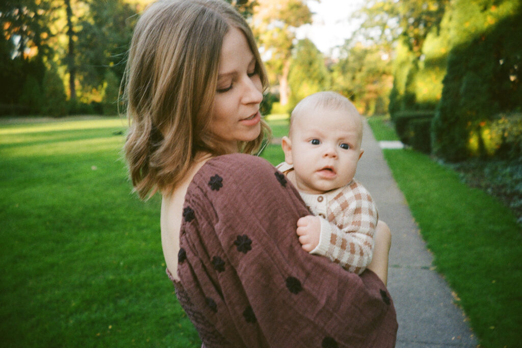 A woman with shoulder-length hair wearing a brown, patterned dress holds a baby in her arms. The baby is dressed in a checkered outfit. They are outside on a paved path surrounded by lush, green grass and trees. The sun casts a warm light.