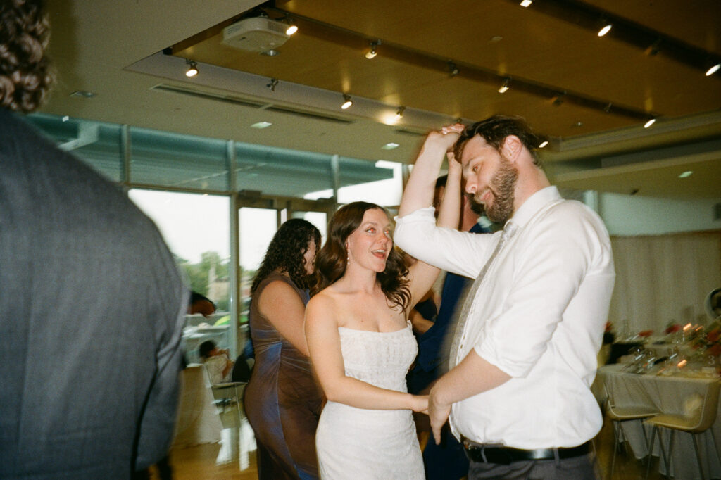 A couple is joyfully dancing in a brightly lit room. The woman, in a white dress, smiles as she dances with a man in a white shirt and tie. Other guests and tables are blurred in the background.