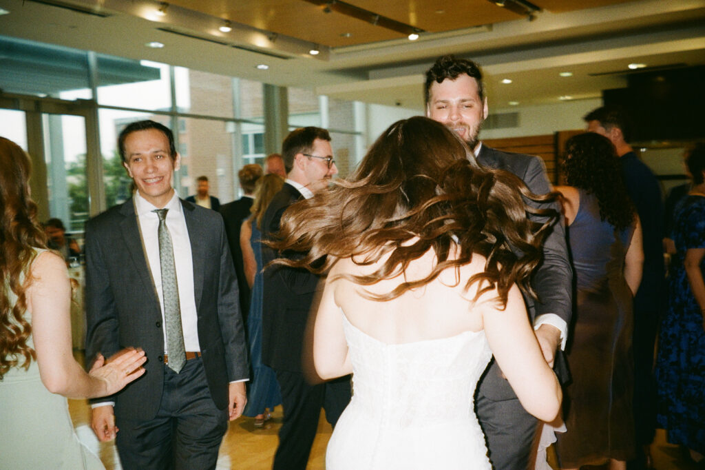 A woman in a white dress is spinning around while dancing with a man in a suit at a gathering. People in formal attire are smiling and engaging in the background. The setting appears to be an indoor event space.