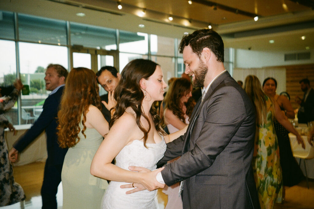 A bride and groom dance closely, smiling in a warmly lit venue. Other guests dance in the background, some laughing and enjoying themselves. The atmosphere is joyful and festive.