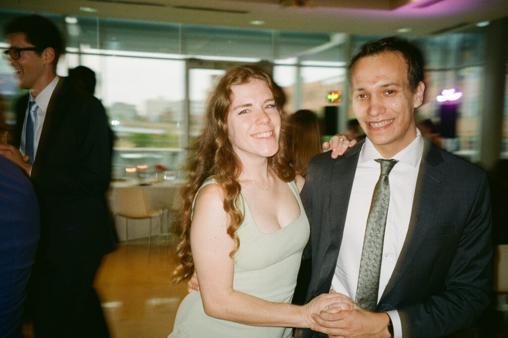 A man and woman are smiling and holding hands at an indoor event. The woman has long brown hair and wears a light green dress, while the man wears a dark suit with a patterned tie. People in the background are mingling in a well-lit room.