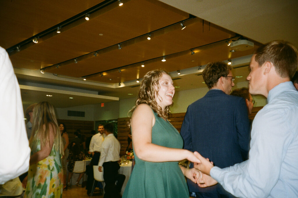 People are dancing happily in a well-lit room with a wooden ceiling. A woman in a green dress and a man in a blue suit are in focus, holding hands. Others in the background are socializing and celebrating.